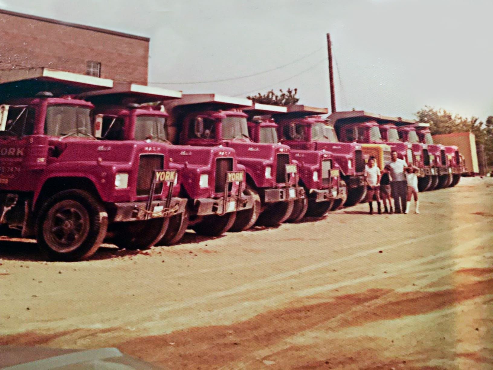 Vintage style photo. Row of red York trucks parked with Santo, York's founder, and his three sons standing in front