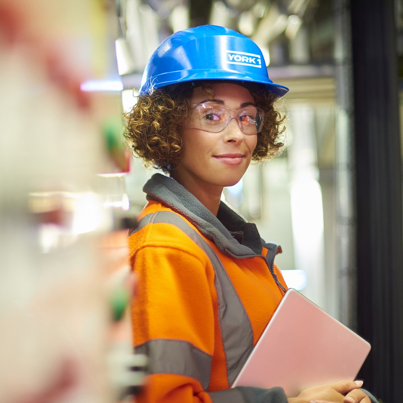 Smiling worker carrying a tablet. Wearing blue York1 hardhat, clear goggles and orange hi-vis jacket.