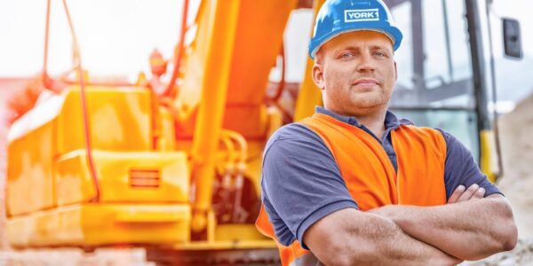 YORK1 worker on a job site, arms crossed, wearing a blue hardhat and orange hi-vis vest. Excavator parked in the background.