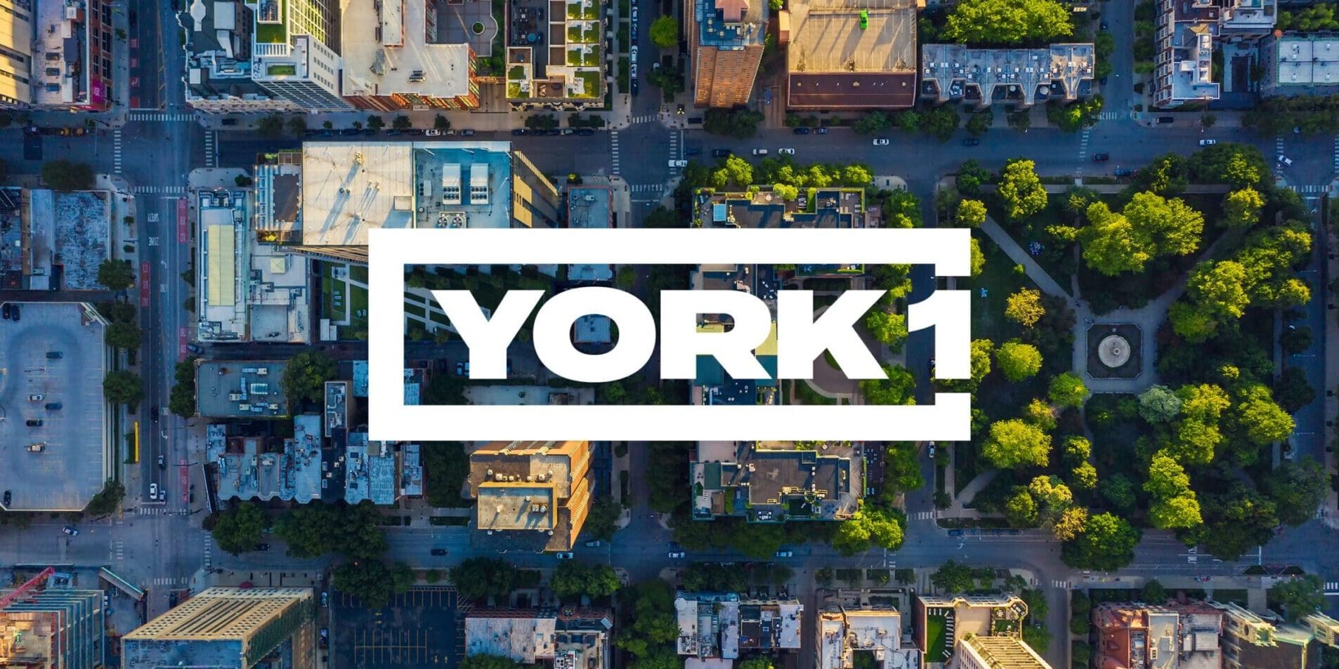 Bird's-eye view of cityscape. Buildings, busy streets, trees, and a park. White York1 logo in the centre.